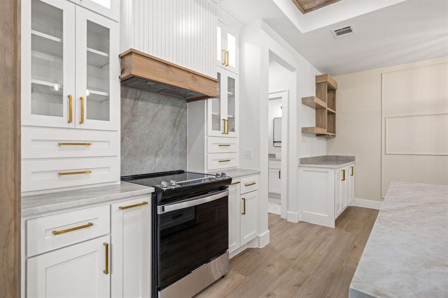 Contemporary kitchen featuring white cabinetry with gold-finish hardware, a built-in stainless steel range, and a wood-finish range hood