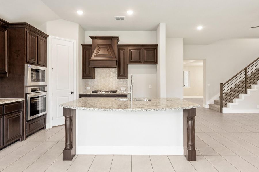 Kitchen with large granite island, dark wood cabinets, custom range hood, and stainless steel appliances on tile floors Kitchen with large granite island, dark wood cabinets, custom range hood, and stainless steel appliances on tile floors