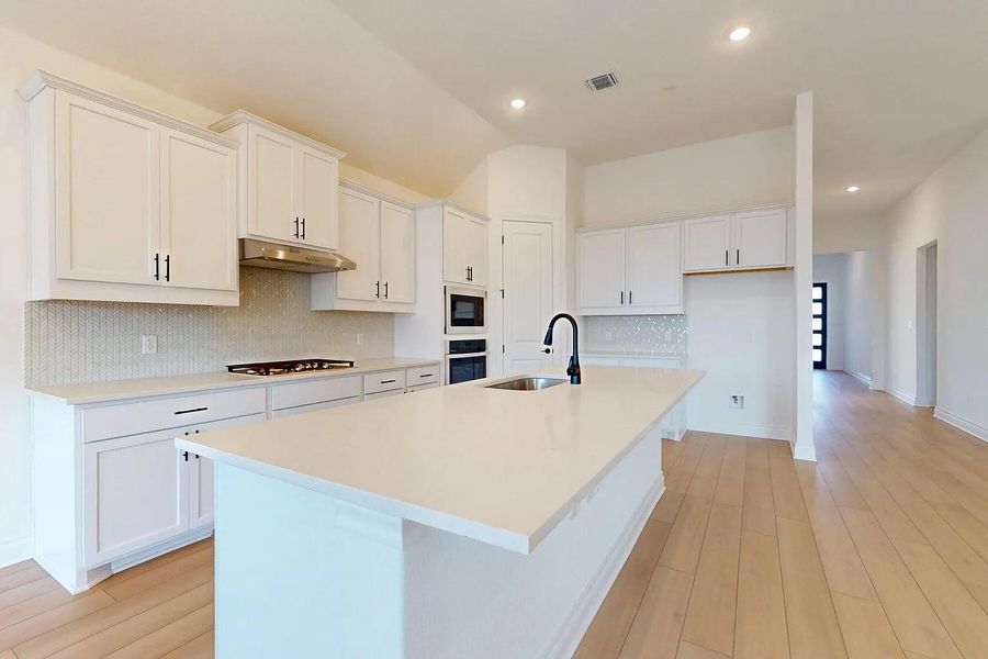 The kitchen features a large island with a sink, white cabinetry with dark hardware, a gas cooktop, and built-in wall ovens