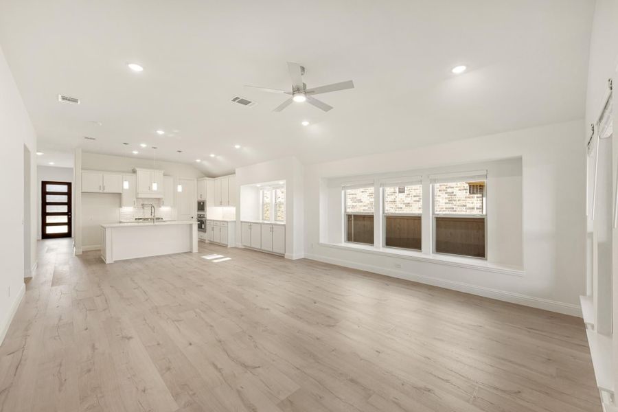 Open-concept living area with light wood floors, ceiling fan, and white kitchen with island in background.