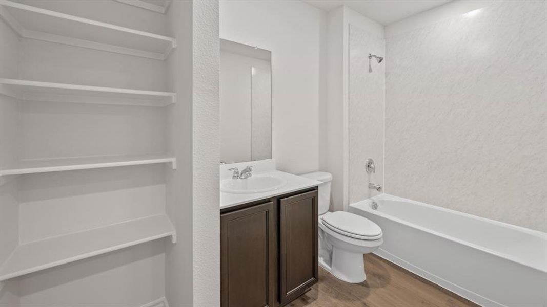 Bathroom with a single vanity, wood-finish flooring, a toilet, and a white tub with an integrated showerhead and textured surround