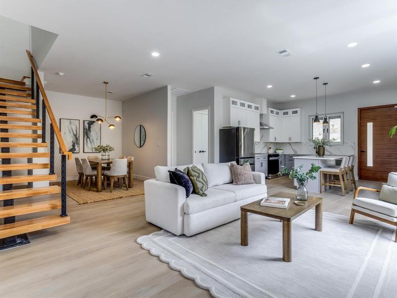 Open-concept living space featuring wood-finish flooring, a modern kitchen with white cabinetry and stainless steel appliances, a dining area with contemporary lighting, and an open-tread staircase with wood and metal railing Open-concept living space featuring wood-finish flooring, a modern kitchen with white cabinetry and stainless steel appliances, a dining area with contemporary lighting, and an open-tread staircase with wood and metal railing