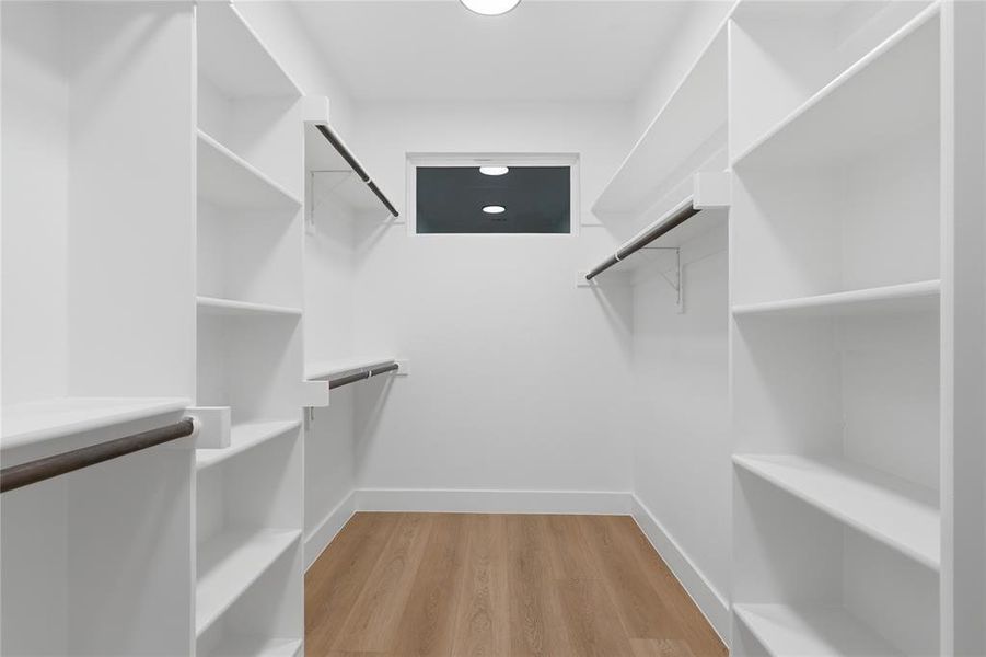 Walk-in closet featuring extensive white shelving, multiple hanging rods, a transom window, wood-finish flooring, and bright overhead lighting Walk-in closet featuring extensive white shelving, multiple hanging rods, a transom window, wood-finish flooring, and bright overhead lighting