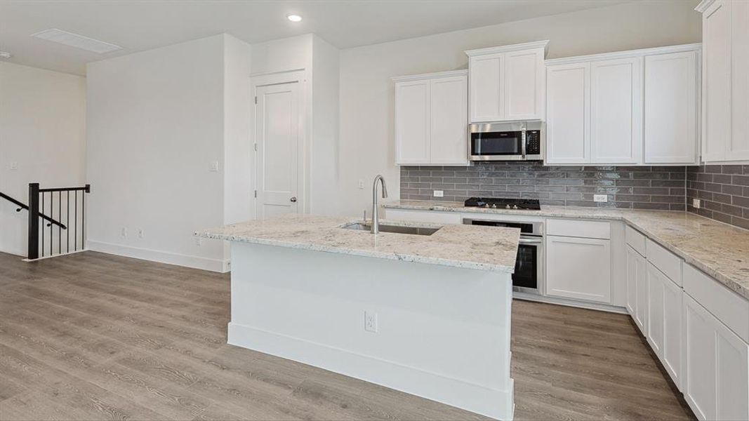 Contemporary kitchen featuring white cabinetry, light granite countertops, a subway tile backsplash, stainless steel appliances, and a central island with an integrated sink