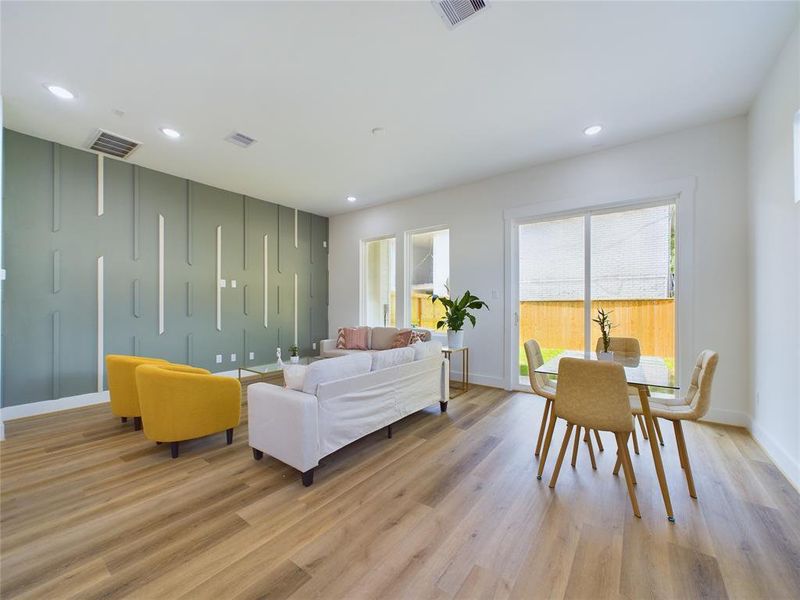 This is a bright, modern open-flow living space with a seamless blend of living, dining, and kitchen areas, featuring light luxury plank floors, white cabinetry, a decorative accent wall. Ample natural light flows throughout.