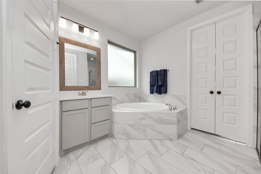 This bathroom features elegant marble tile flooring, a spacious corner bathtub, and a modern vanity with a large mirror. The room is bright and airy, with frosted windows for privacy and ample storage behind double doors.