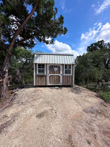 A charming rustic shed nestled amongst natural landscaping under a blue sky.