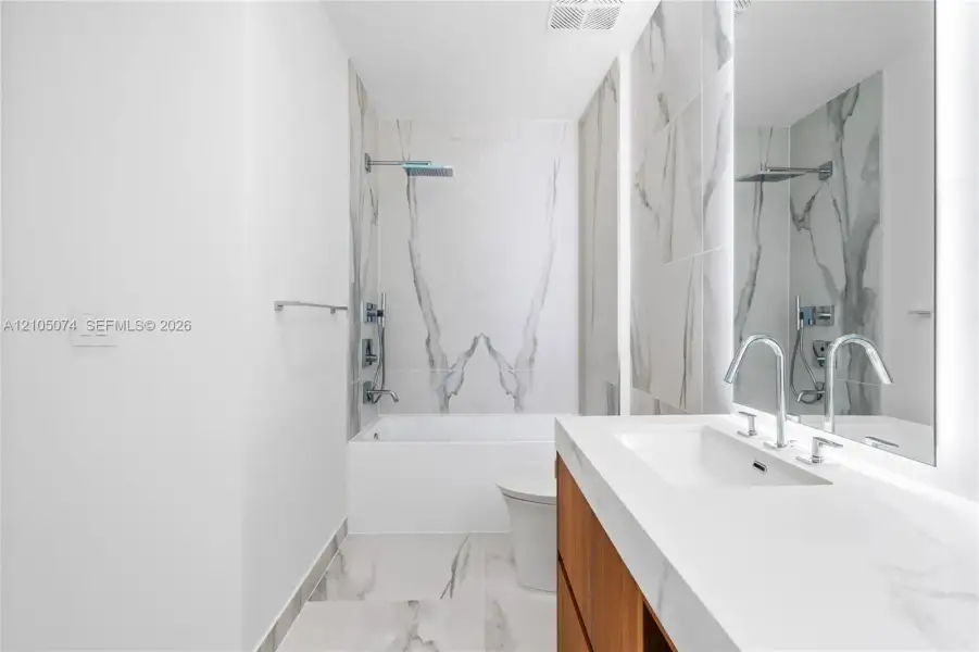 Bathroom featuring a wood-finish vanity with a stone countertop, large-format tile flooring, a bathtub-shower combination with wall-to-ceiling stone-patterned tile, and an oversized mirror