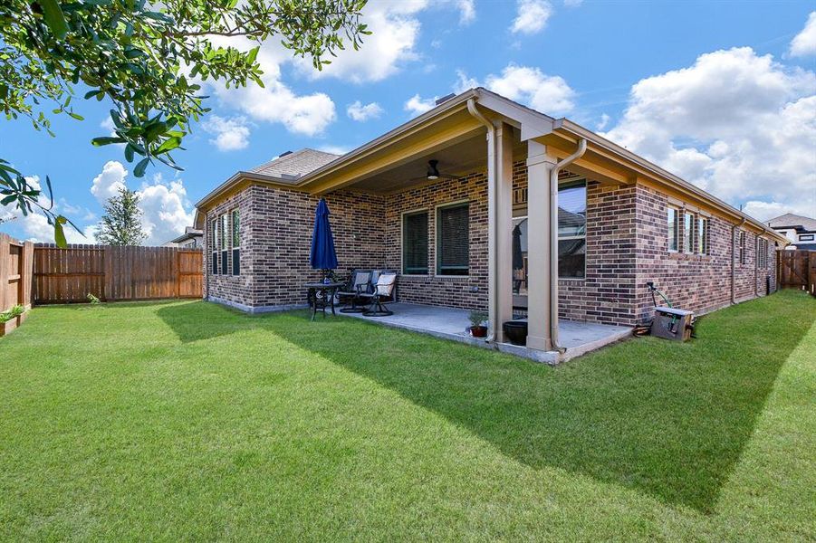 This photo showcases a well-maintained backyard with a spacious covered patio attached to a brick house. The yard is enclosed with a wooden fence, providing privacy, and features a small dining set perfect for outdoor gatherings. The lawn is green and inviting, ideal for relaxation or play.