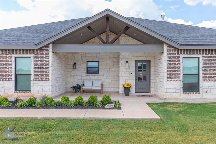 Property entrance with a porch, a shingled roof, stone siding, and a yard Property entrance with a porch, a shingled roof, stone siding, and a yard