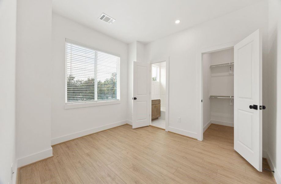 Bright secondary bedroom with high ceilings, recessed lighting, large window, warm wood-look flooring, and direct access to a closet and nearby bath. The neutral finishes create a calm, versatile space that works beautifully as a bedroom, guest room, nursery, or private home office.
