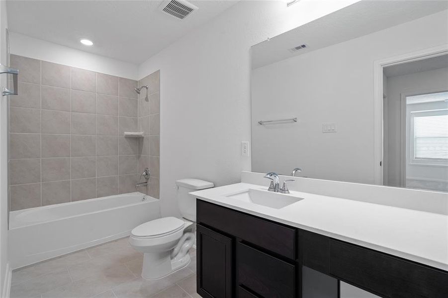 Secondary bath features tile flooring, bath/shower combo with tile surround, dark stained wood cabinets, beautiful light countertops, mirror, dark, sleek fixtures and modern finishes.