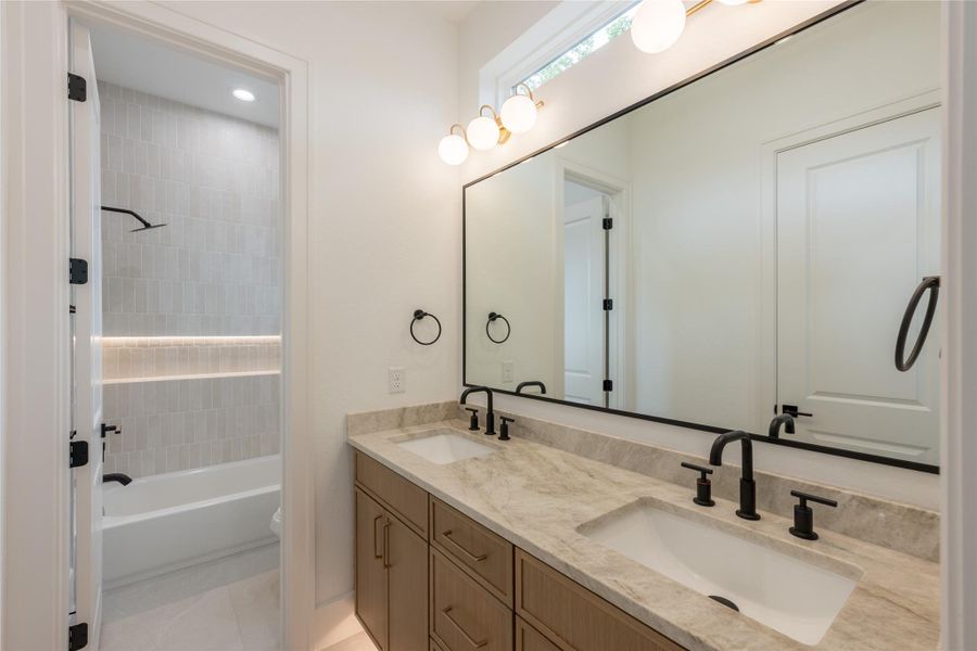 A streamlined secondary bath where wood cabinetry and stone counters create warmth complemented by dual vanities designed for everyday ease.