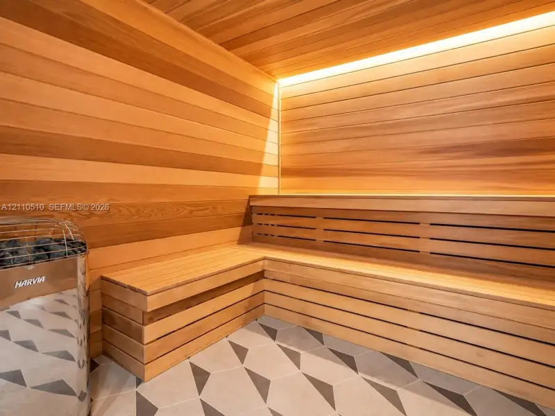 Sauna featuring wood-plank walls and ceiling, integrated multi-level bench seating, a chrome-finish Harvia heater, and geometric-pattern floor tile with recessed linear accent lighting Sauna featuring wood-plank walls and ceiling, integrated multi-level bench seating, a chrome-finish Harvia heater, and geometric-pattern floor tile with recessed linear accent lighting