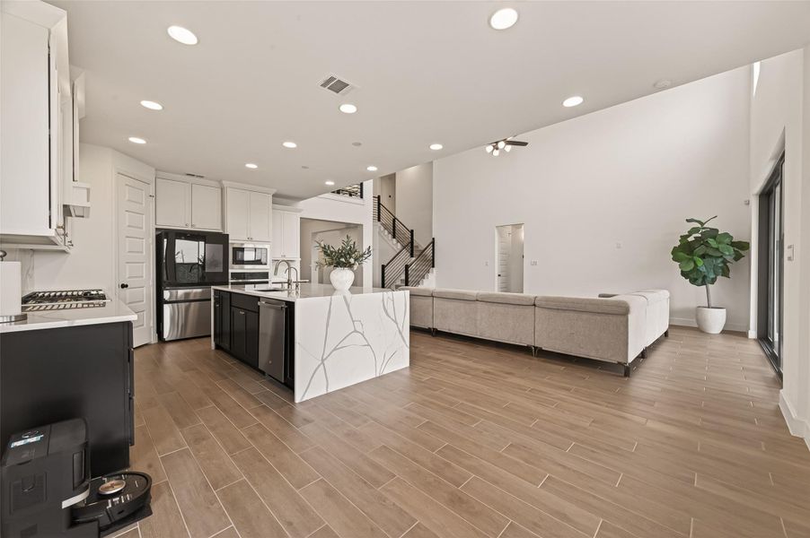 This spacious, modern open-concept living area features a sleek kitchen with a large island, ample cabinetry, and stainless steel appliances. It flows into a bright living room with high ceilings, wood-style flooring, and large windows, creating a welcoming and airy atmosphere.