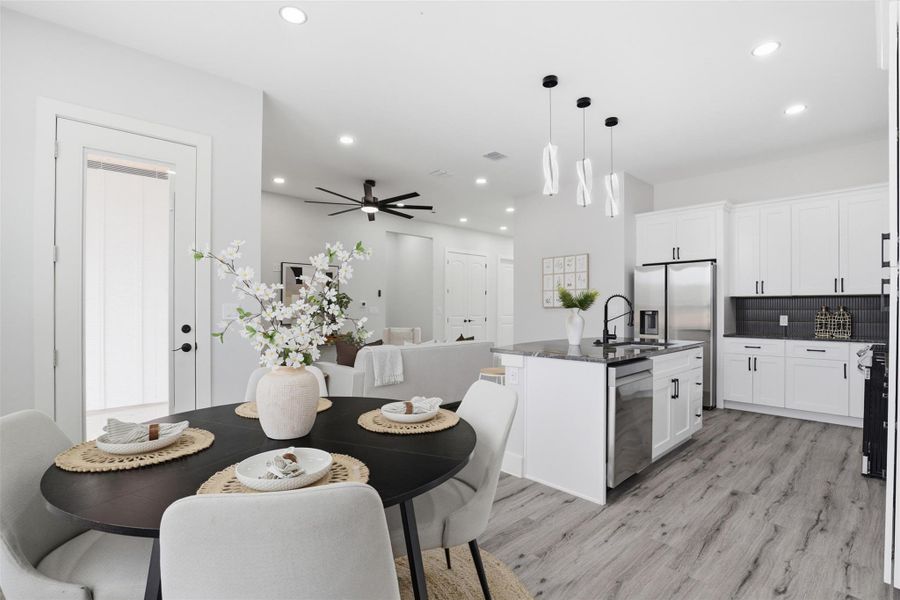 Modern kitchen and dining area featuring wood-finish flooring, white cabinetry, a dark tiled backsplash, stainless steel appliances, and an island with pendant lighting