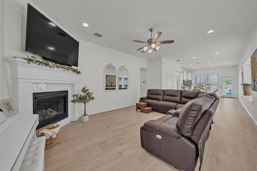 Open-concept living space featuring a fireplace with a white mantel, recessed lighting, a ceiling fan with integrated lighting, wood-finish flooring, and multiple windows providing natural light Open-concept living space featuring a fireplace with a white mantel, recessed lighting, a ceiling fan with integrated lighting, wood-finish flooring, and multiple windows providing natural light
