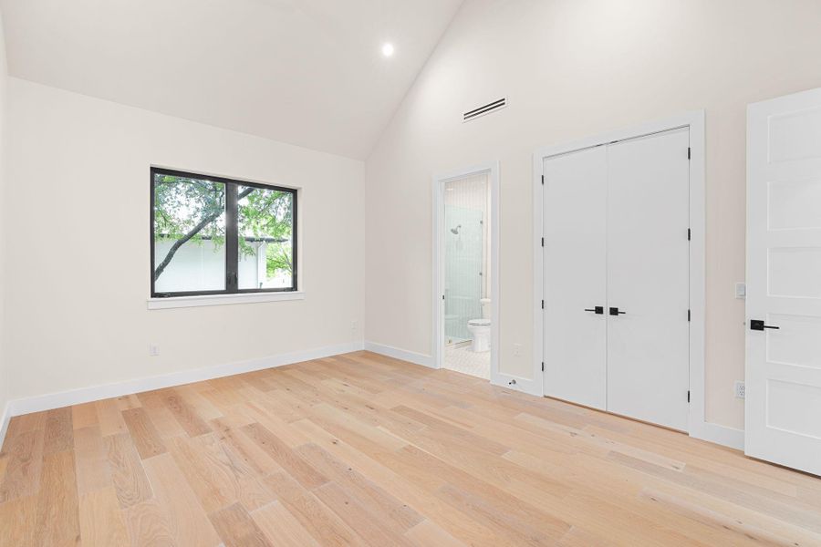 Room featuring light wood-finish flooring, a vaulted ceiling, a black-framed window, and a door to a bathroom with a glass-enclosed shower