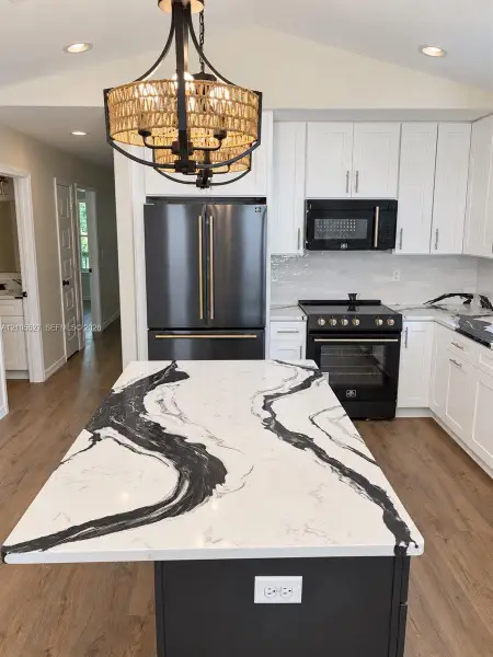 Kitchen featuring white upper cabinetry, dark lower island cabinetry, dark stainless steel appliances, wood-finish flooring, and a decorative chandelier Kitchen featuring white upper cabinetry, dark lower island cabinetry, dark stainless steel appliances, wood-finish flooring, and a decorative chandelier