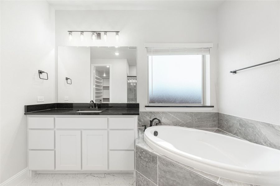 Bathroom with white vanity, dark countertop, large mirror, and soaking tub with gray tile surround