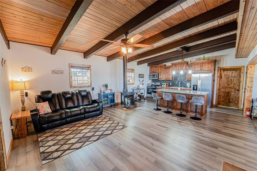 Living room with ceiling fan, a wood stove, light wood-style floors, vaulted ceiling with beams, and wooden ceiling Living room with ceiling fan, a wood stove, light wood-style floors, vaulted ceiling with beams, and wooden ceiling