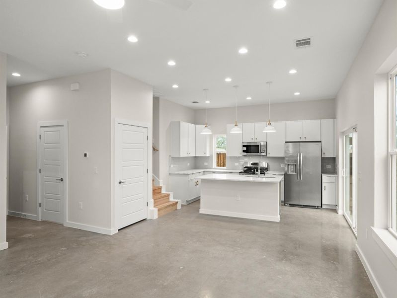 Open-concept living space featuring concrete flooring, recessed lighting, a modern kitchen island with pendant lighting, stainless steel appliances, and abundant natural light from large windows