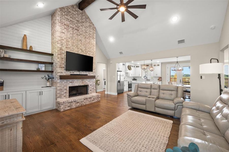 Living room featuring a fireplace, brick wall, ceiling fan with notable chandelier, and high vaulted ceiling