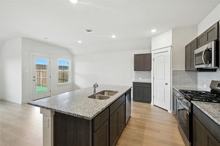 Kitchen featuring stainless steel appliances, backsplash, light stone countertops, dark brown cabinetry, and light wood finished floors Kitchen featuring stainless steel appliances, backsplash, light stone countertops, dark brown cabinetry, and light wood finished floors