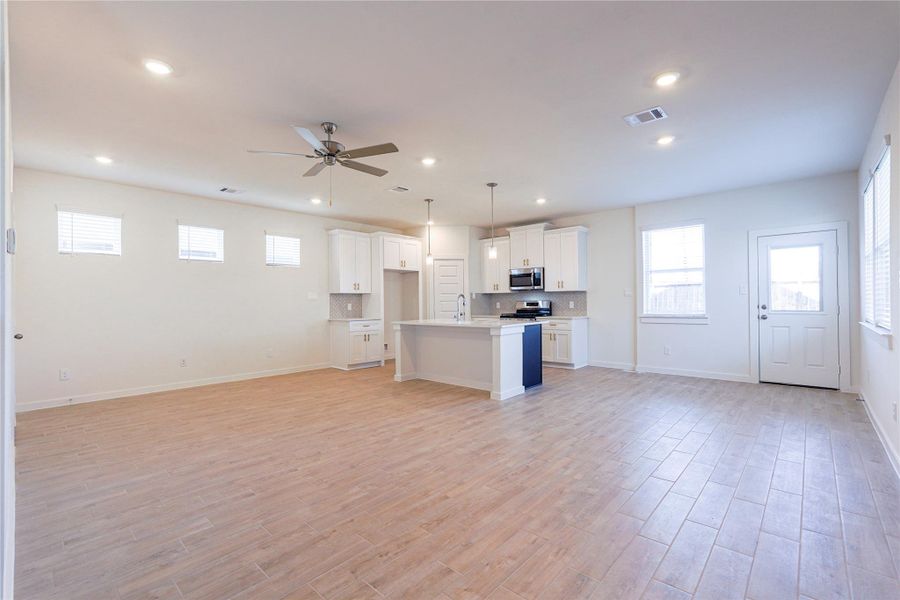 This open-concept living space features modern hardwood flooring, recessed lighting, and a ceiling fan. The kitchen boasts white cabinetry, stainless steel appliances, and a central island with pendant lighting. Ample natural light comes through multiple windows, enhancing the bright and airy feel.