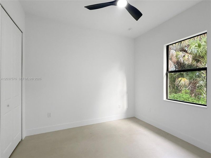 Neutral interior space featuring a ceiling fan with integrated lighting, a window with exterior foliage, white walls, and a light-colored floor