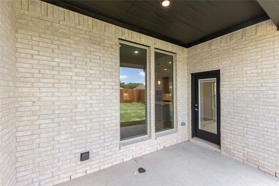 Covered back patio with light brick exterior, sliding glass door, and black entry door overlooking fenced backyard.