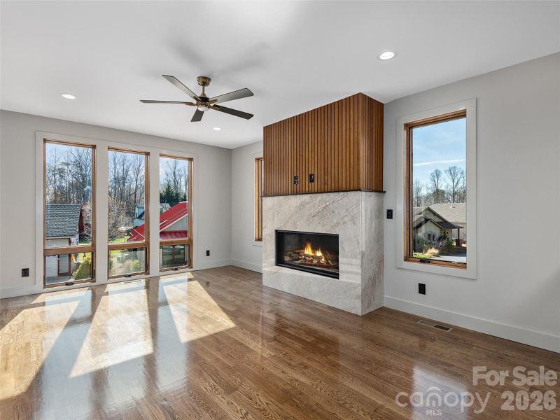 Living room filled with natural light, featuring oak hardwood floors, expansive windows, and a vented gas fireplace with granite surround and custom wood accent detail.