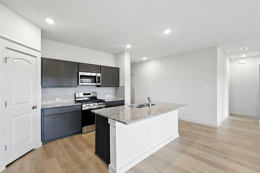 Kitchen with tasteful backsplash, appliances with stainless steel finishes, a center island with sink, light stone countertops, and light wood-style floors Kitchen with tasteful backsplash, appliances with stainless steel finishes, a center island with sink, light stone countertops, and light wood-style floors