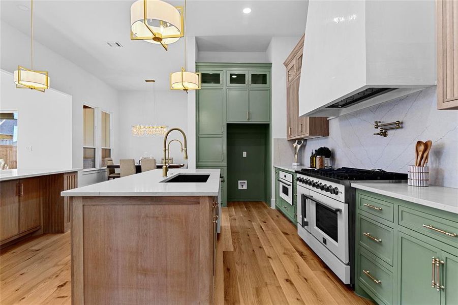 Kitchen featuring white appliances, custom exhaust hood, light quartz counters, quartz backsplash, and green custom cabinetry with a 48inch stove and a farm sink. Vent hood is in a Venetian plaster. Kitchen featuring white appliances, custom exhaust hood, light quartz counters, quartz backsplash, and green custom cabinetry with a 48inch stove and a farm sink. Vent hood is in a Venetian plaster.