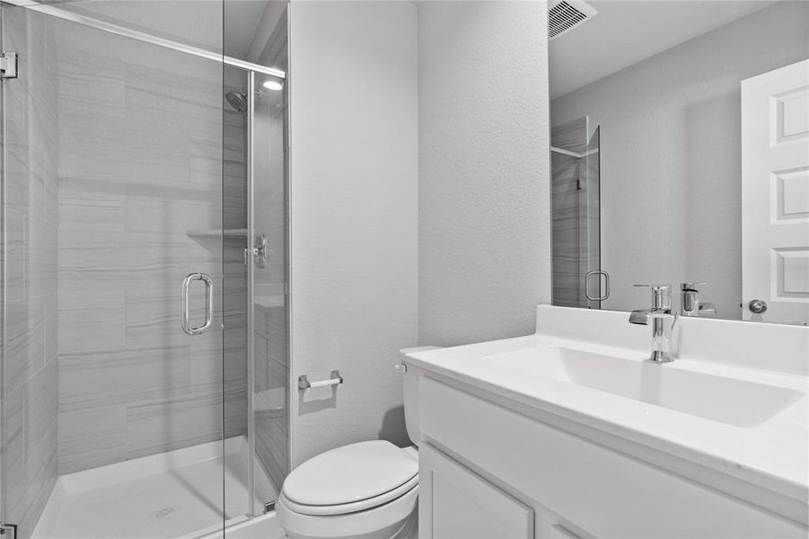 This private bath features tile flooring, shower with tile surround, light stained wood cabinets, beautiful light countertops, mirror, dark, sleek fixtures and modern finishes.