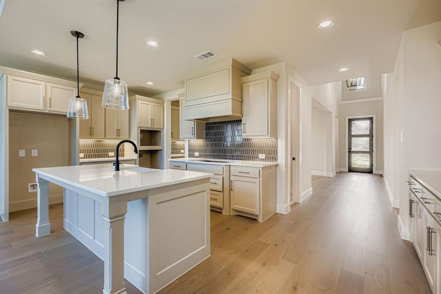 Kitchen featuring backsplash, pendant lighting, light wood-style floors, a center island with sink, and light stone countertops