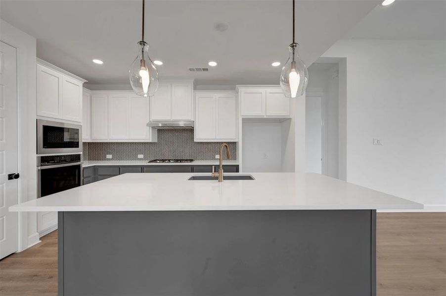 Modern kitchen featuring a large island with a white countertop and dark grey base, white upper cabinetry, grey lower cabinetry, stainless steel appliances, and two pendant lights