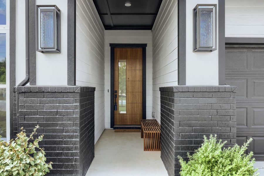 The entry welcomes you with a striking wood-paneled door framed by modern sconces and dark brick pillars.