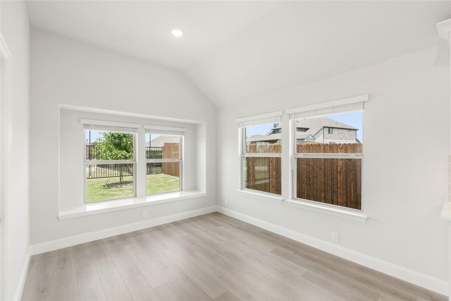 Empty room with vaulted ceiling, light wood floors, and multiple windows overlooking a fenced backyard Empty room with vaulted ceiling, light wood floors, and multiple windows overlooking a fenced backyard