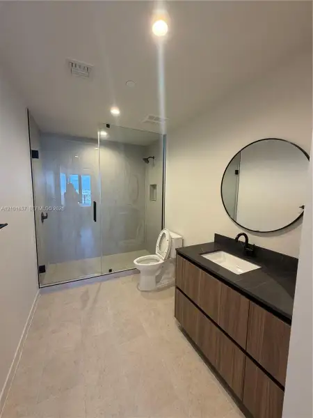 Floating vanity featuring wood-finish cabinetry, dark countertop, and integrated rectangular sink, paired with a frameless glass-enclosed shower, matte black fixtures, and a circular wall-mounted mirror