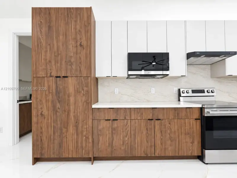 Kitchen featuring two-tone cabinetry with wood-finish lower units and floor-to-ceiling pantry, white upper cabinets, stone-surfaced backsplash, white countertops, and stainless steel appliances