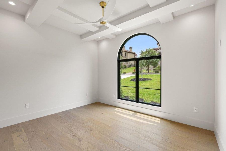 Room featuring an arched black-frame window, light wood-finish flooring, white walls, and a multi-tiered ceiling with a gold-accented ceiling fan and recessed lighting