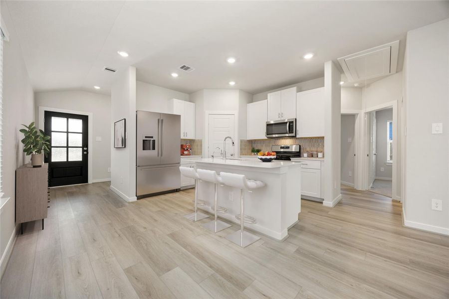 This modern kitchen features crisp white cabinetry, quartz countertops, and stainless steel appliances. A large center island provides ample workspace and casual seating, while the open layout and warm-toned vinyl plank flooring create a bright, inviting atmosphere.