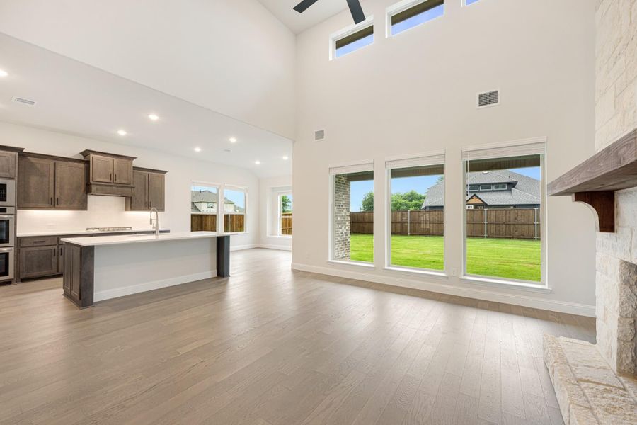 Open-concept living area with hardwood floors, high ceilings, stone fireplace, and kitchen with dark cabinets and center island Open-concept living area with hardwood floors, high ceilings, stone fireplace, and kitchen with dark cabinets and center island