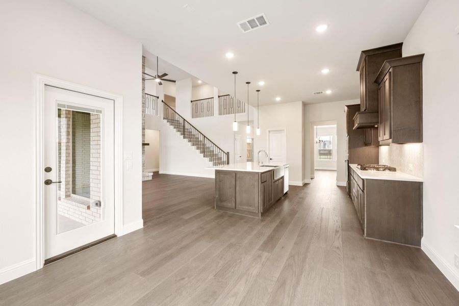 Open-concept kitchen and living area with dark wood cabinets, center island, hardwood floors, and staircase with upper loft railing.