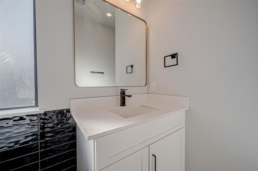 This modern bathroom features a sleek white vanity with a stylish black faucet, a large mirror, and contemporary lighting. The contrasting black tile adds a touch of elegance.