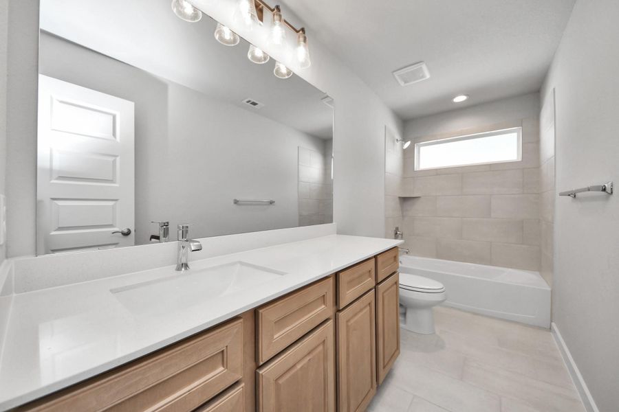 Secondary bath features tile flooring, bath/shower combo with tile surround, light stained wood cabinets, beautiful light countertop.