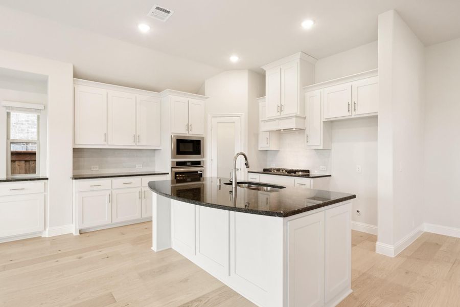 Kitchen with white cabinets, dark granite island with sink, stainless steel appliances, and light hardwood floors