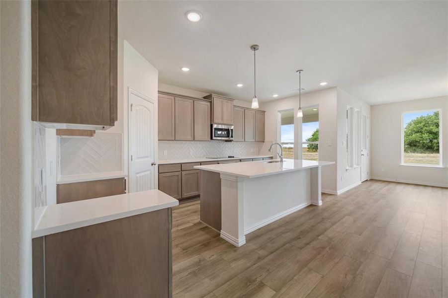 Kitchen with tasteful backsplash, hanging light fixtures, an island with sink, light wood-style floors, and stainless steel microwave