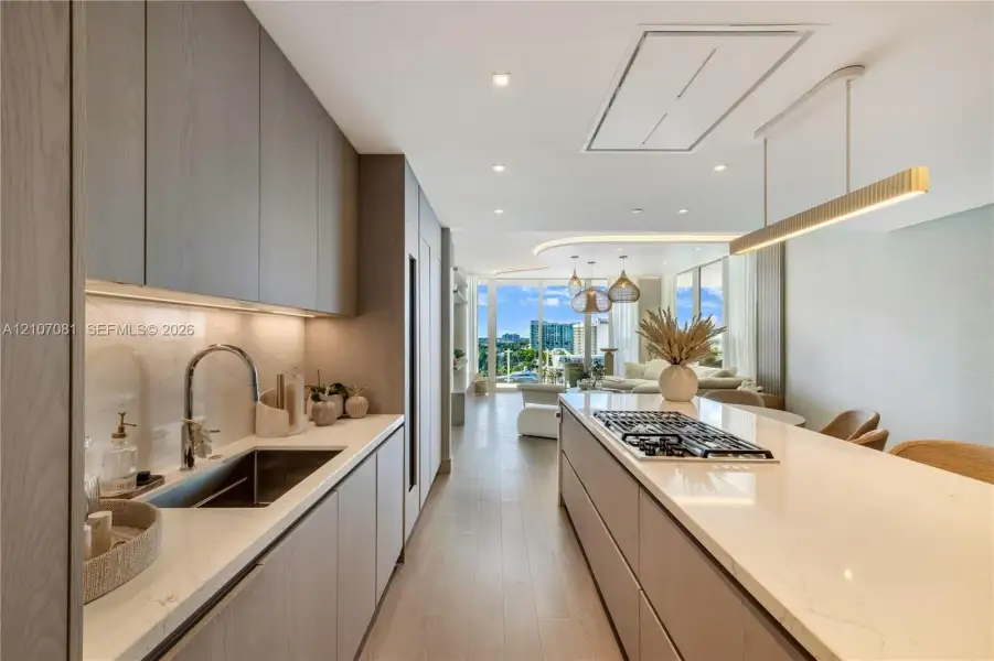 Contemporary kitchen featuring flat-panel cabinetry, light-toned stone countertops, a large center island with an integrated gas cooktop, wood-finish flooring, and recessed ceiling lighting Contemporary kitchen featuring flat-panel cabinetry, light-toned stone countertops, a large center island with an integrated gas cooktop, wood-finish flooring, and recessed ceiling lighting